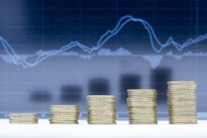 Pilhas de moedas de altura crescente, com gráficos financeiros em segundo plano, simbolizando o crescimento da poupança e a evolução das taxas de juro.